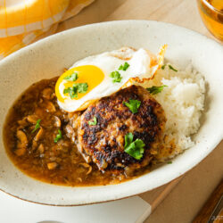 A plate of white rice topped with a hamburger patty, brown mushroom gravy, and a sunny-side-up egg, garnished with parsley. A yellow checkered napkin and a glass of tea are beside the plate.