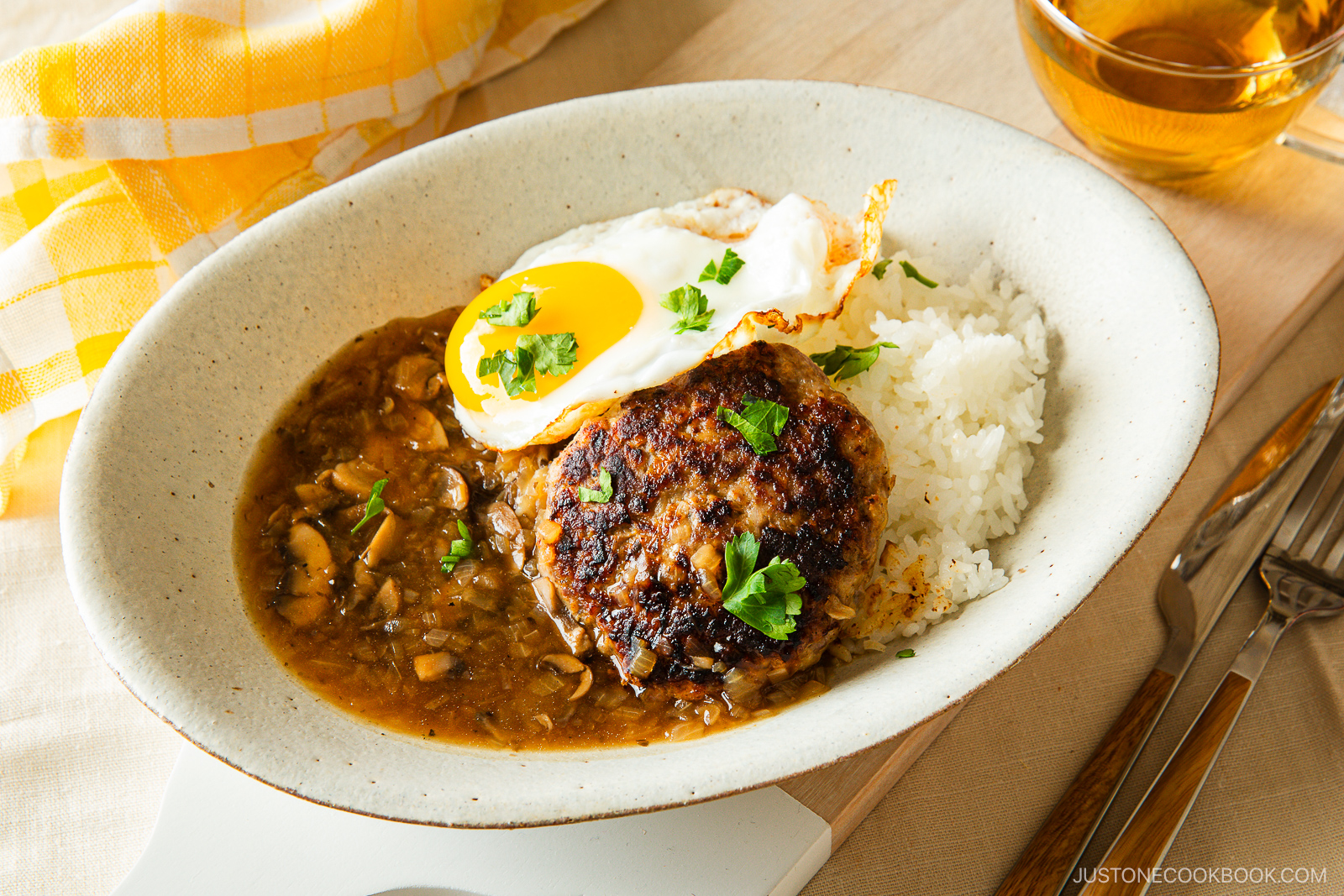 A plate of white rice topped with a hamburger patty, brown mushroom gravy, and a sunny-side-up egg, garnished with parsley. A yellow checkered napkin and a glass of tea are beside the plate.