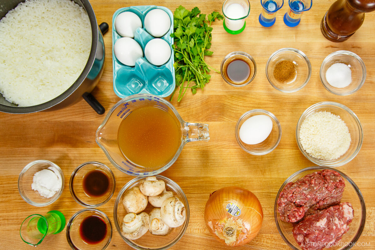 Top-down view of various ingredients on a wooden surface, including cooked rice, eggs, ground meat, onion, mushrooms, fresh herbs, chicken broth, seasonings, and small bowls of sauces and spices.