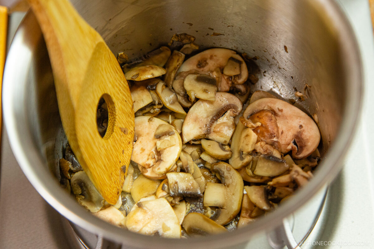 Sliced mushrooms being sautéed in a stainless steel pot, stirred with a wooden spatula.