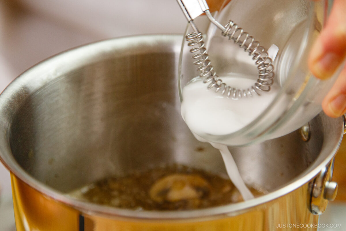 A hand pours a white liquid mixture from a small glass bowl into a pot of simmering liquid on the stove, using a metal whisk.