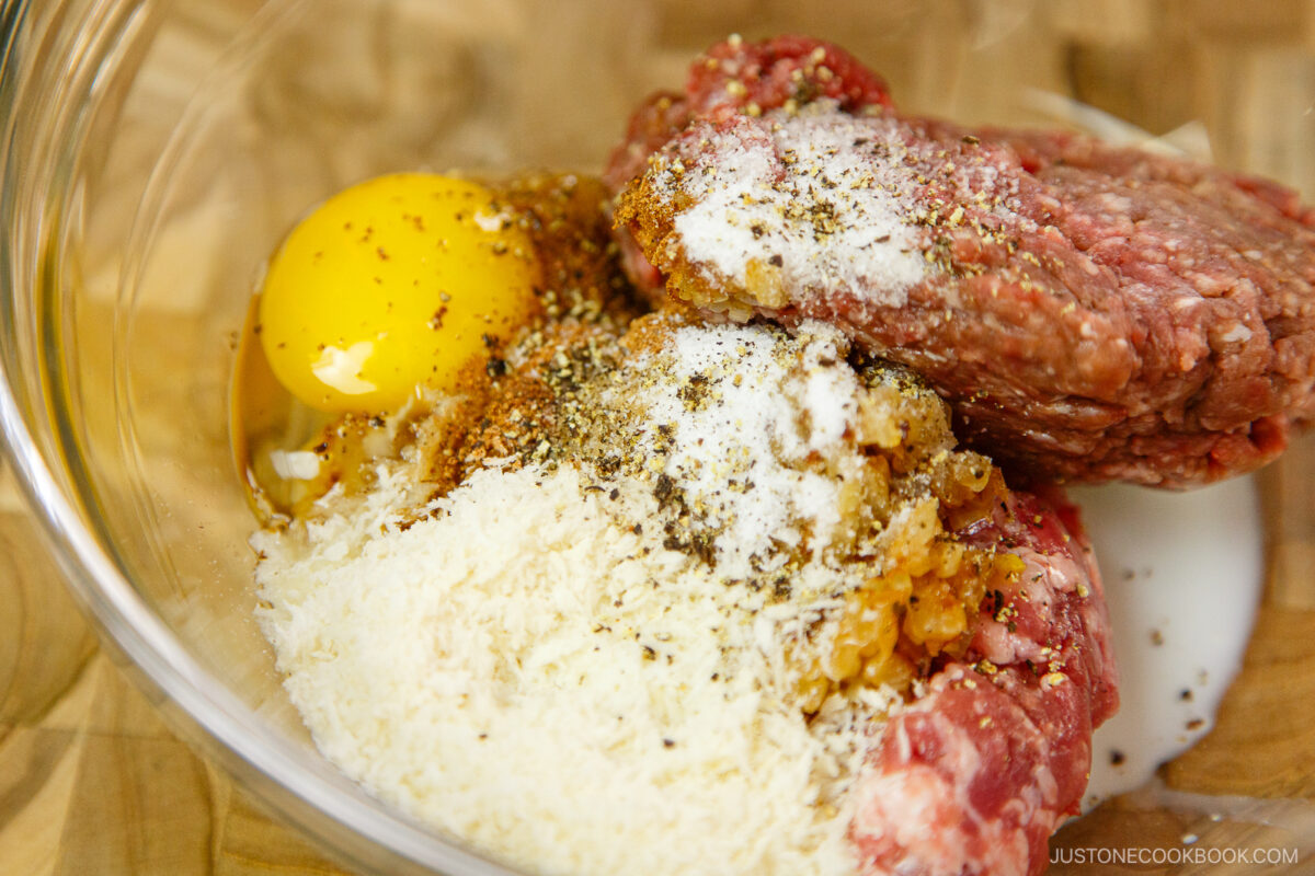 A glass bowl contains raw ground beef, a cracked egg, grated cheese, seasonings, and a small amount of milk, ready to be mixed for a recipe.