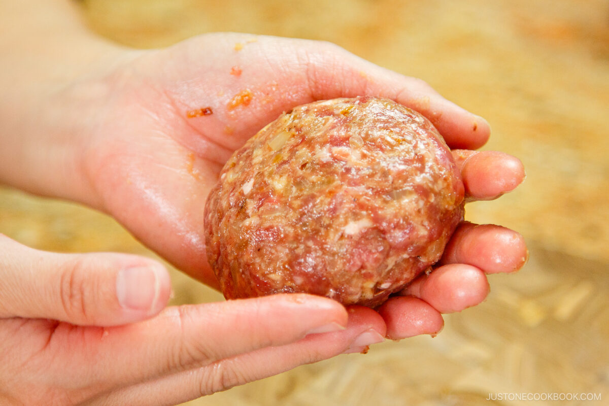 A pair of hands shaping a ball of raw ground meat mixture, preparing it for cooking. The mixture contains visible bits of meat and fat.