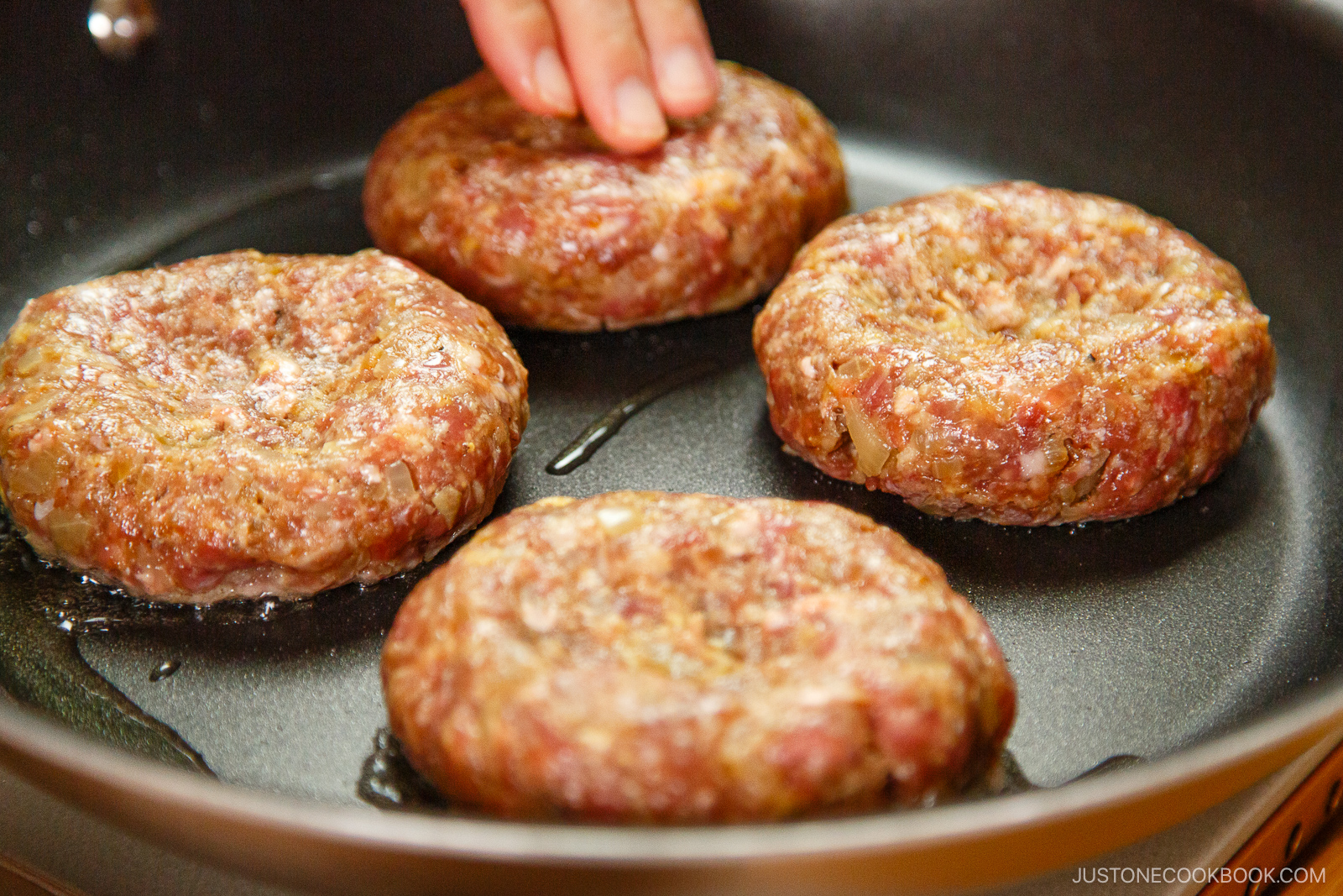 Four raw hamburger patties are being arranged in a frying pan by a hand, ready to be cooked. The patties appear fresh with visible minced meat and seasonings.