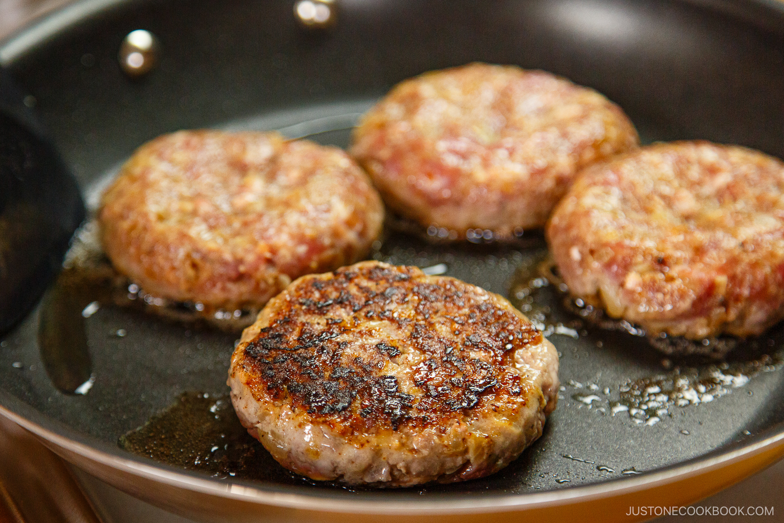 Four hamburger patties are sizzling in a frying pan. One patty in the foreground is browned and cooked, while the others are still cooking. A bit of oil is visible in the pan.