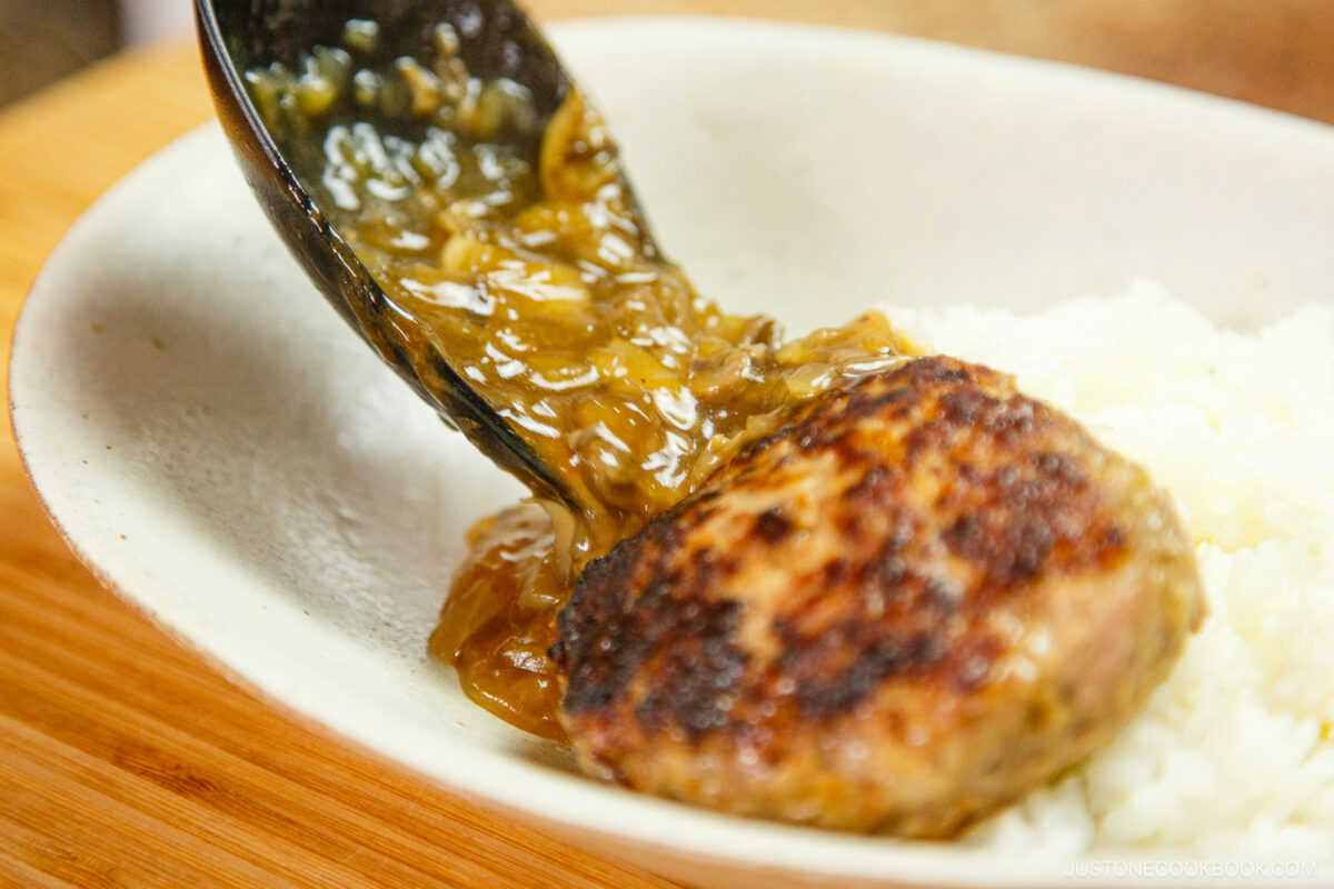 A close-up of a browned hamburger steak topped with onion gravy, served with white rice in a bowl. A spoon is pouring the gravy over the steak.