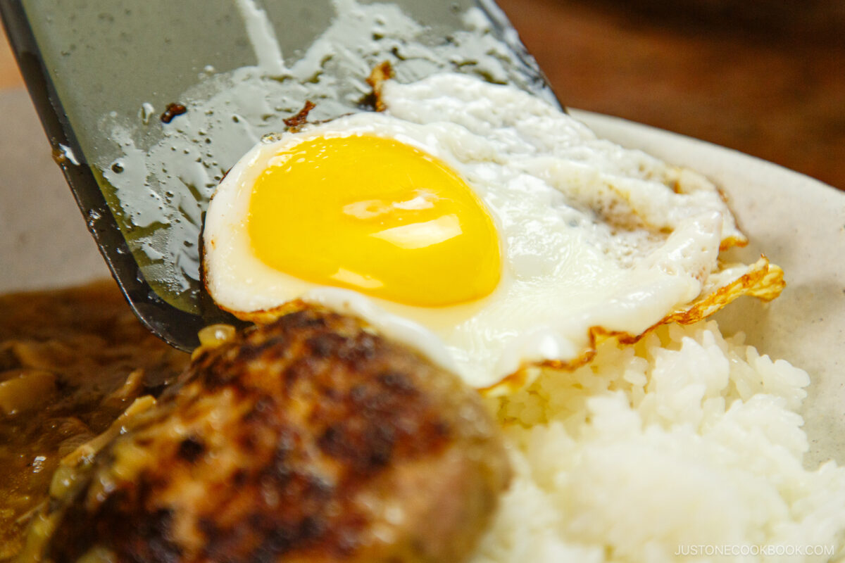 A spatula lifts a sunny-side-up egg onto a plate with white rice and a hamburger patty, forming part of a loco moco dish.