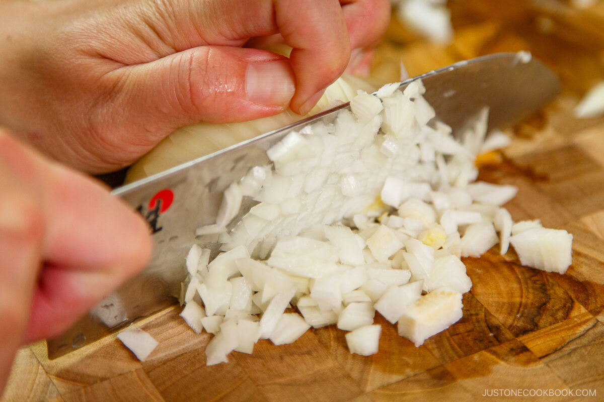 Close-up of hands finely dicing a white onion with a large knife on a wooden cutting board.