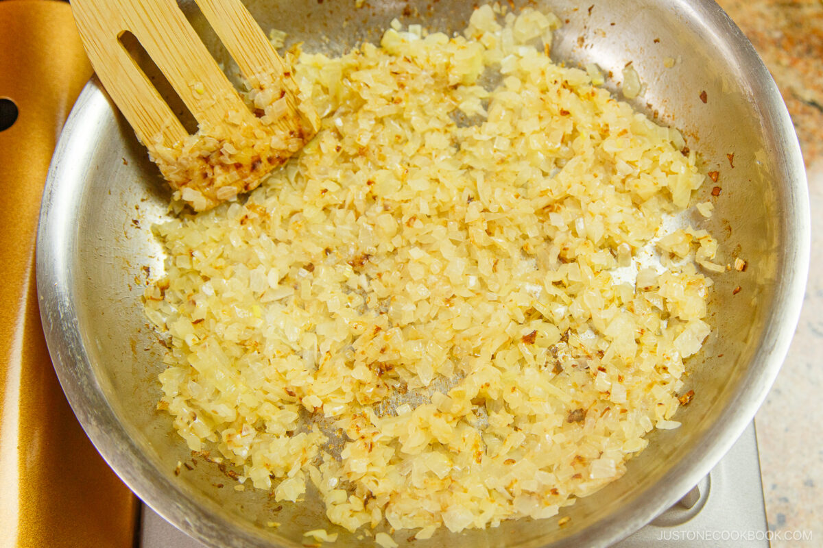 Chopped onions being sautéed in a stainless steel pan, turning golden brown, with a wooden spatula stirring them.