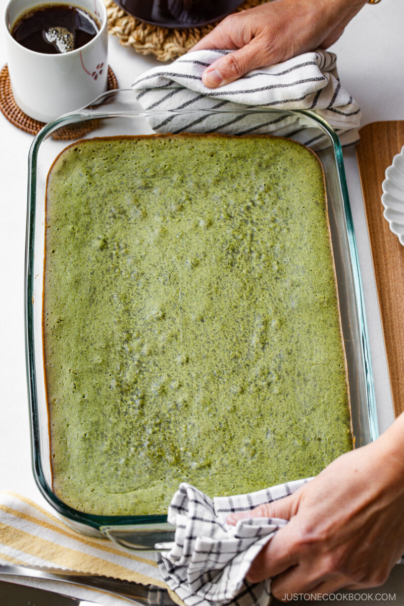A person holds a glass baking dish filled with a green sheet cake, using kitchen towels for grip. A cup of coffee and plates are visible nearby on a white tabletop.