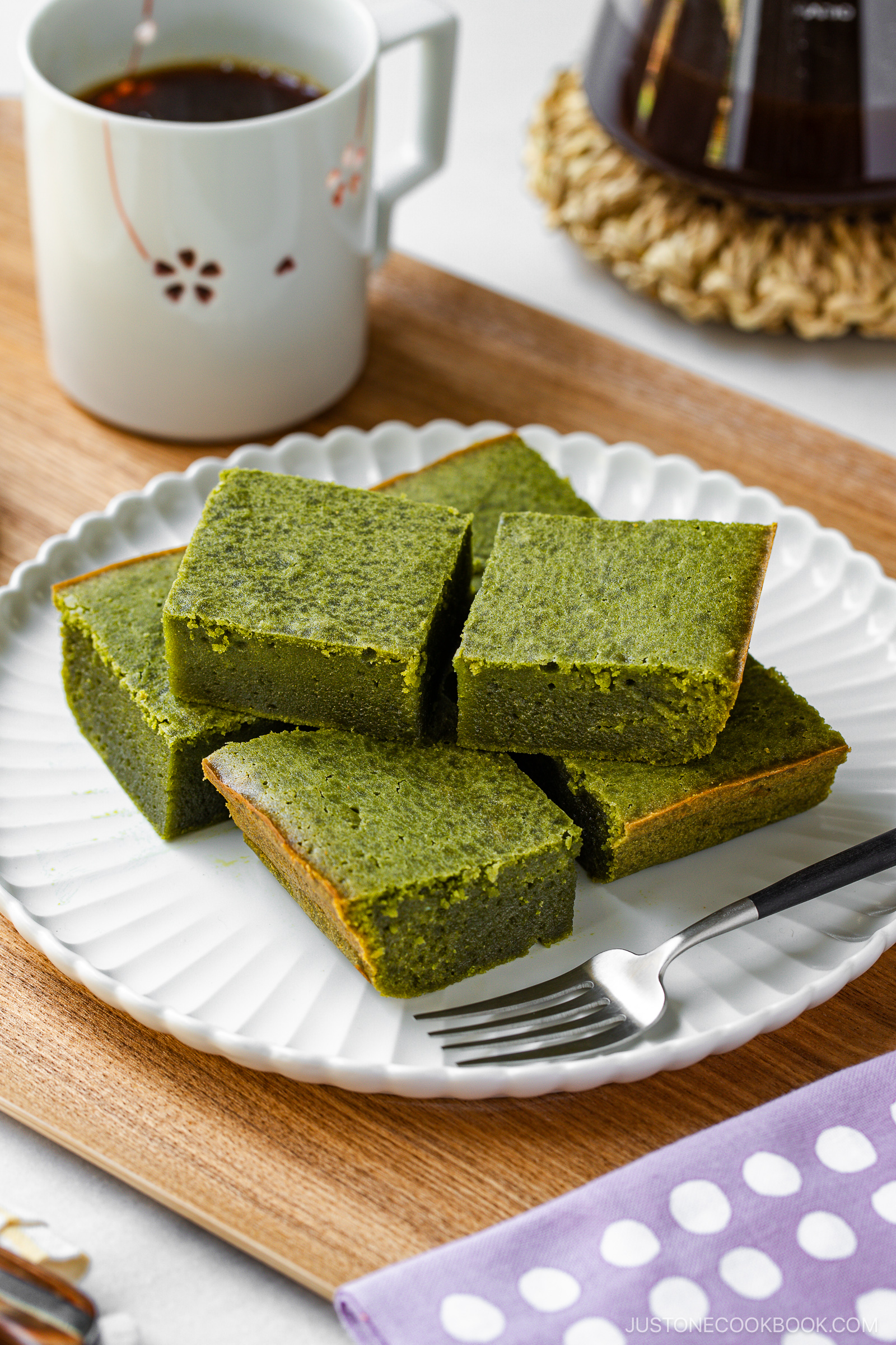 Six square pieces of green matcha cake are arranged on a white plate with a fork beside them. A cup of black coffee and a coffeepot sit in the background on a wooden surface.