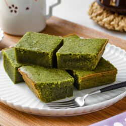 A white plate with six square pieces of green matcha brownies is placed on a wooden tray. A black-handled fork rests beside the brownies, and a white mug sits in the background.