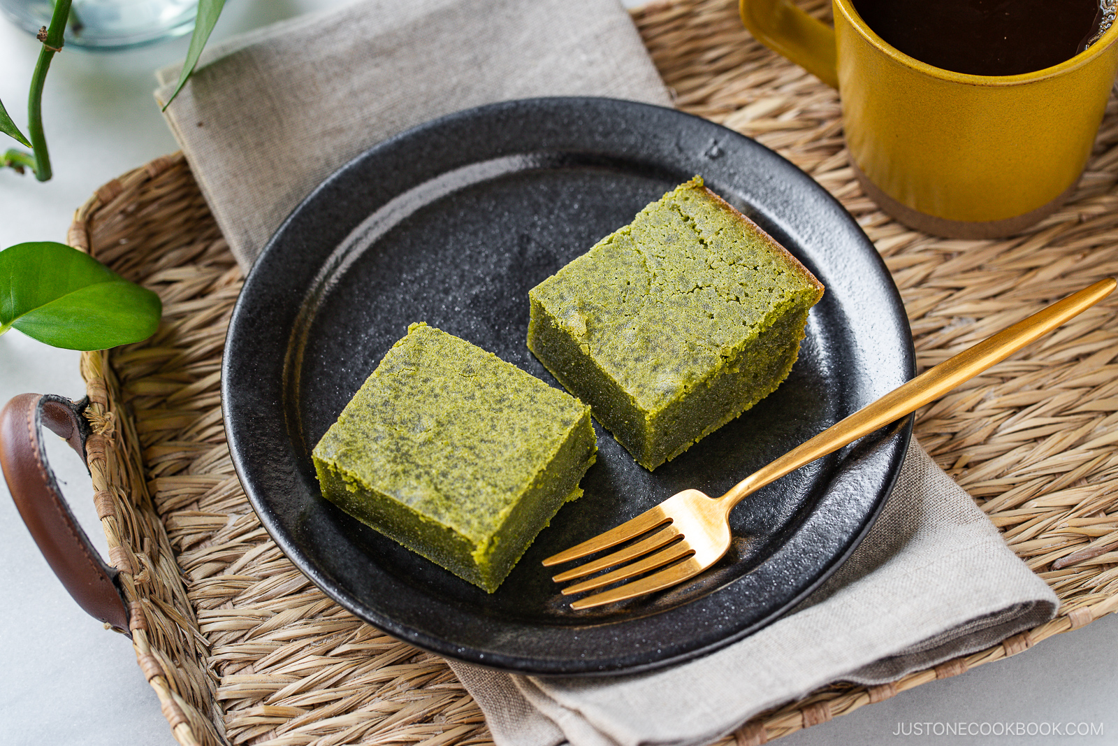 Two green matcha brownie squares on a black plate with a gold fork, placed on a beige napkin atop a woven tray. A cup of coffee and green plant are partially visible in the background.