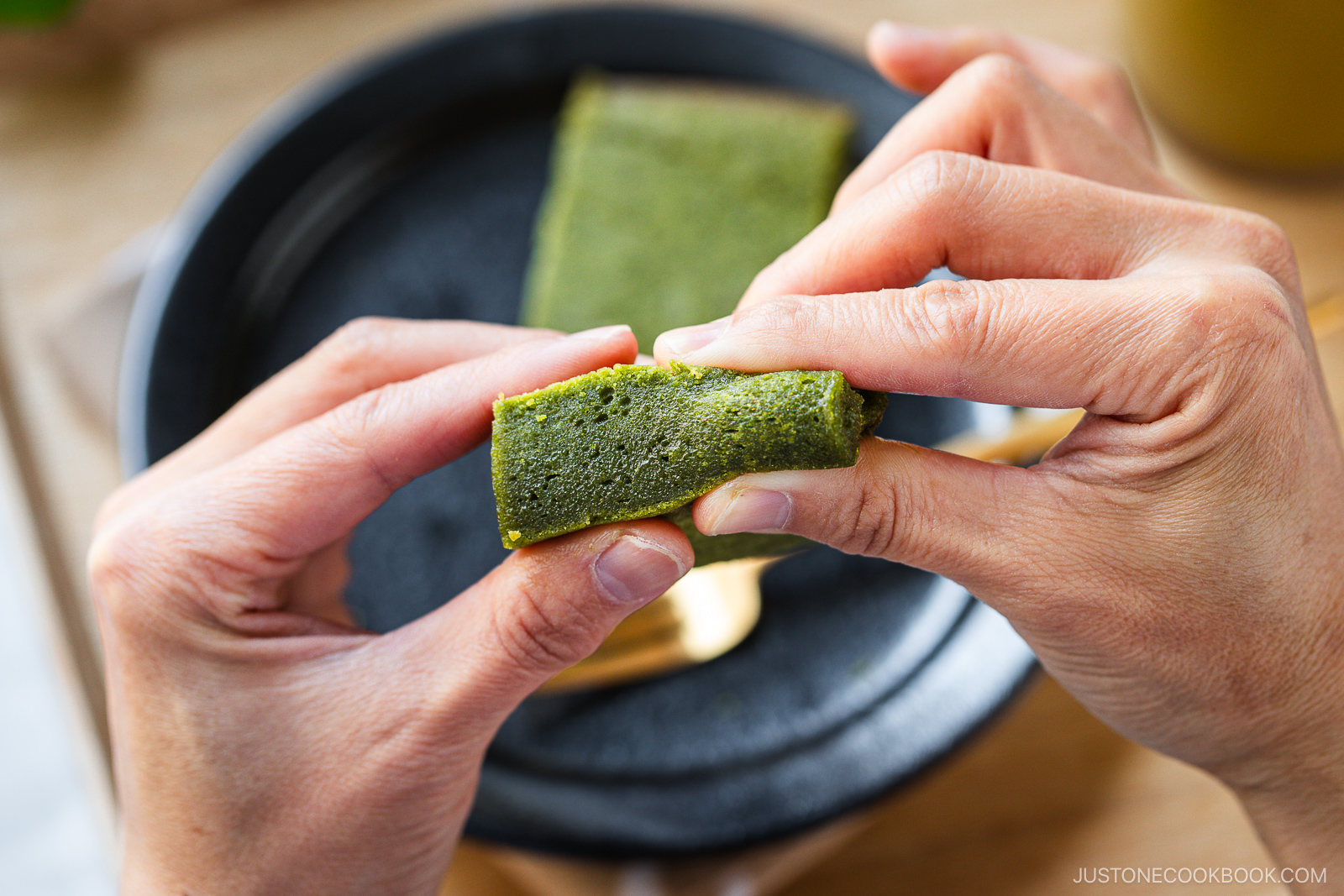Two hands gently pull apart a rectangular slice of green matcha cake over a black plate. The cake looks soft and moist.