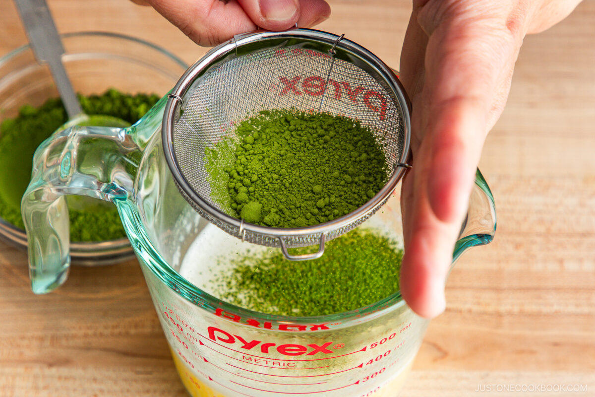 A hand holds a fine mesh strainer above a glass measuring cup, sifting bright green matcha powder into a liquid. A small bowl of more matcha powder sits in the background on a wooden surface.