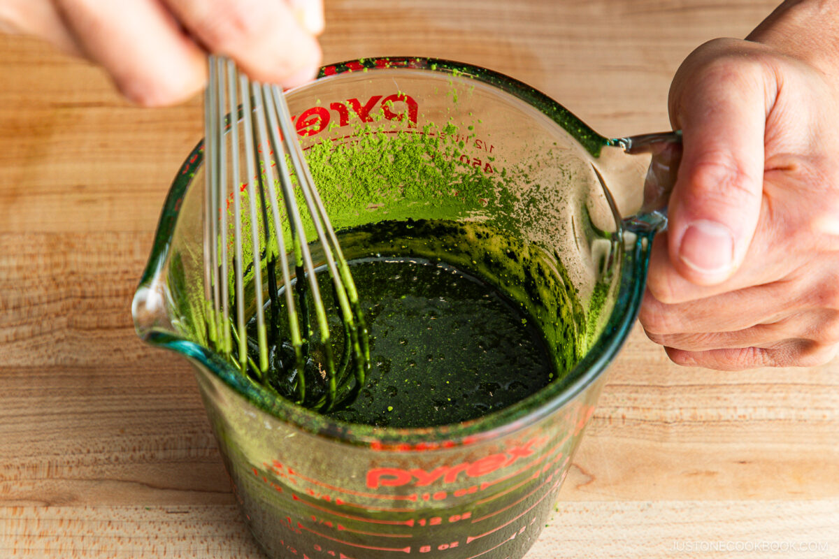 A person whisks a green liquid in a glass Pyrex measuring cup on a wooden surface.