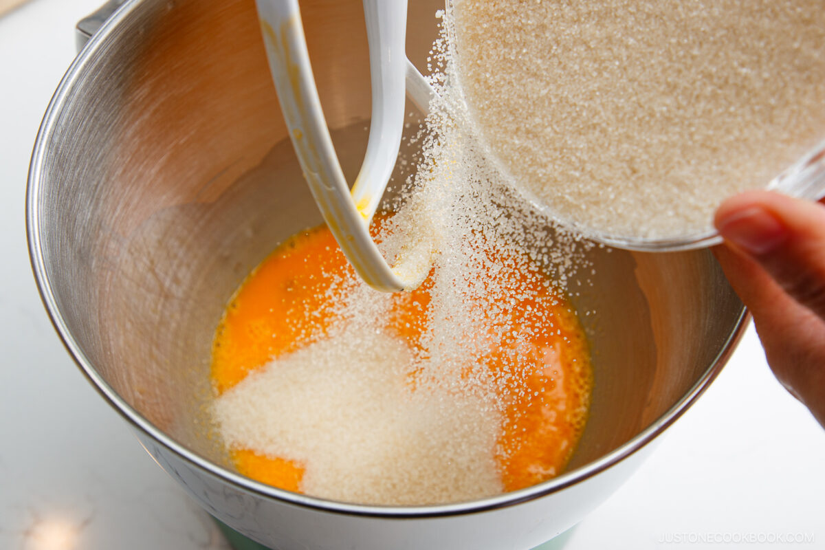 A hand pours granulated sugar from a bowl into a stand mixer bowl containing beaten eggs, preparing ingredients for baking.