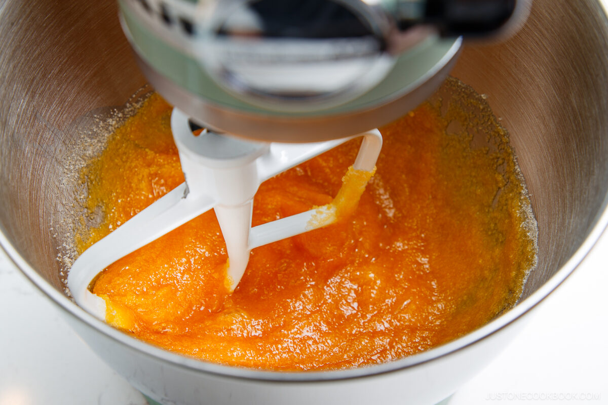 A stand mixer with a paddle attachment is blending bright orange ingredients in a metal bowl, likely sugar and egg yolks being creamed together.