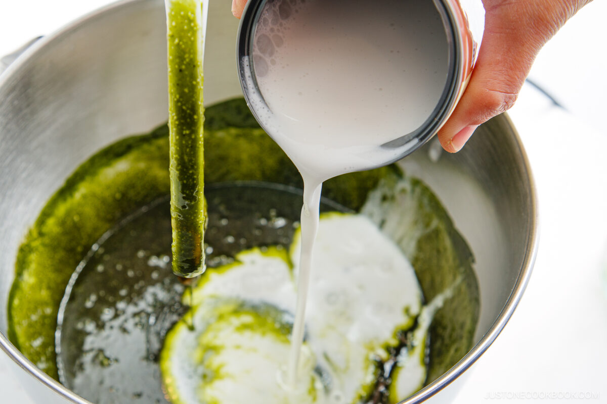 A hand pours coconut milk from a small bowl into a metal mixing bowl containing a thick green matcha mixture.