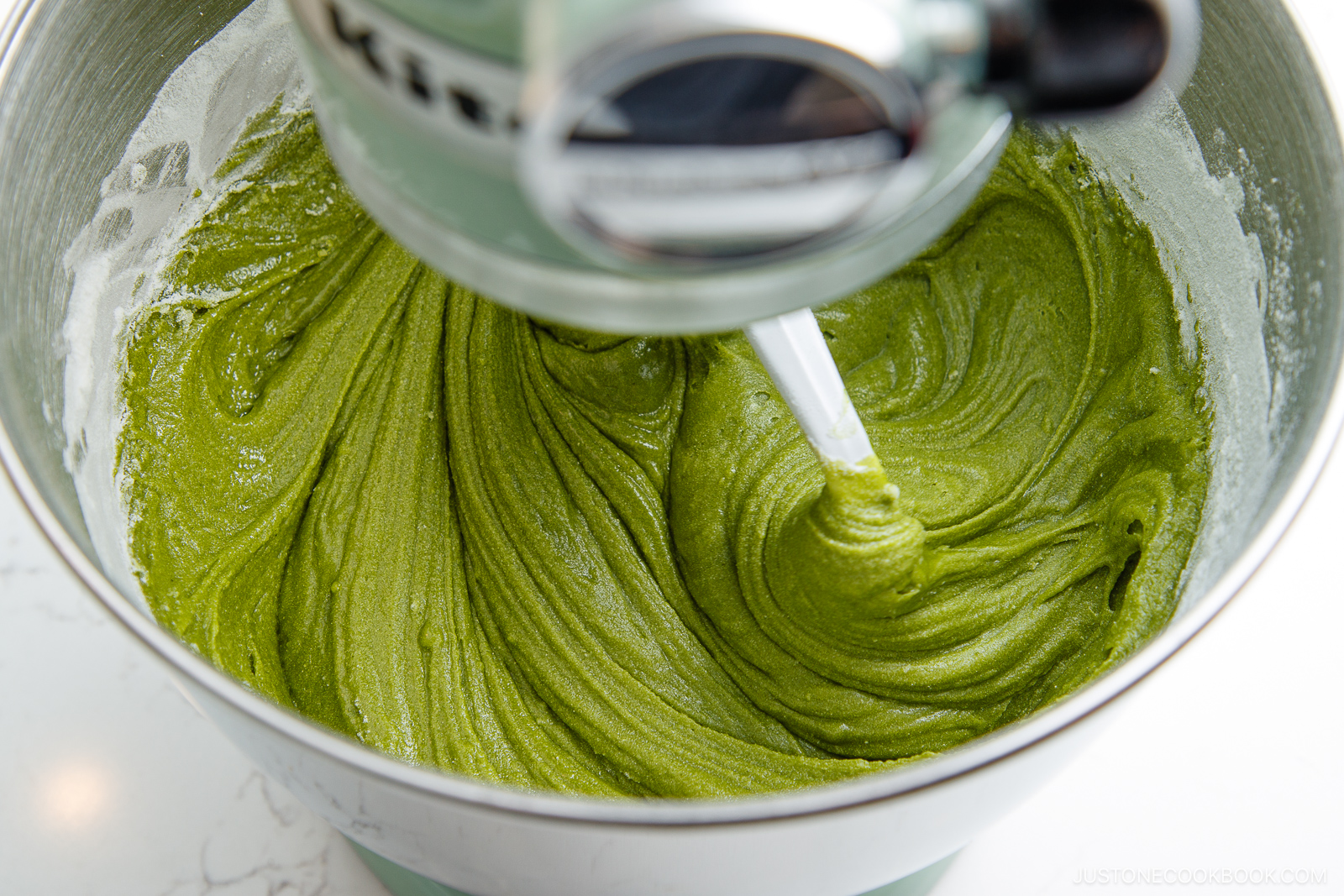 A stand mixer bowl filled with smooth, bright green matcha batter being mixed by a paddle attachment. The sides of the bowl have traces of flour.
