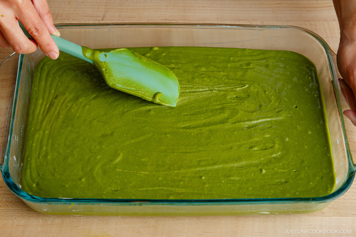 A person uses a green spatula to spread green matcha cake batter evenly in a rectangular glass baking dish on a wooden surface.