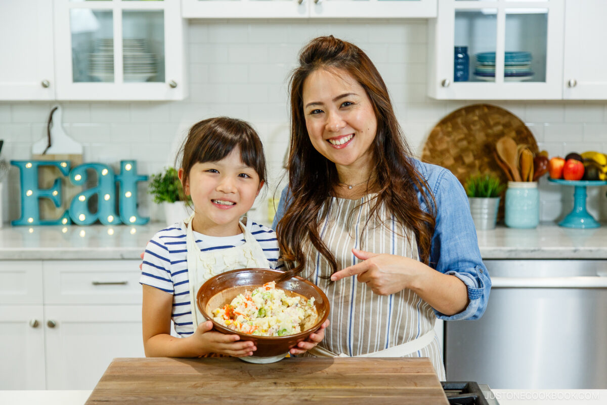 A smiling woman and a young girl stand in a bright kitchen, holding a bowl of salad together. The woman is pointing to the bowl. Both are wearing aprons, and kitchen decor is visible in the background.