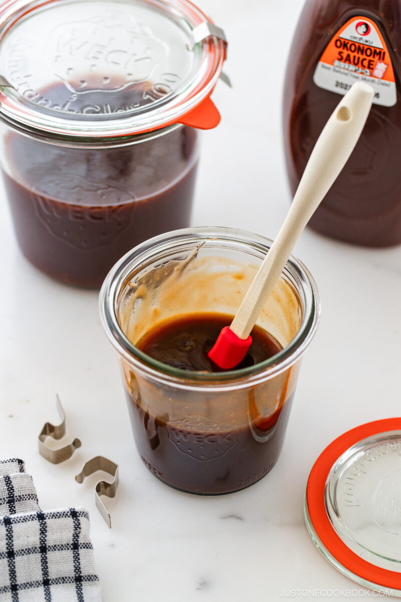 A glass jar filled with dark brown okonomiyaki sauce and a red spatula sits on a white surface, with another sealed jar, a sauce bottle, metal clips, and a checkered cloth nearby.