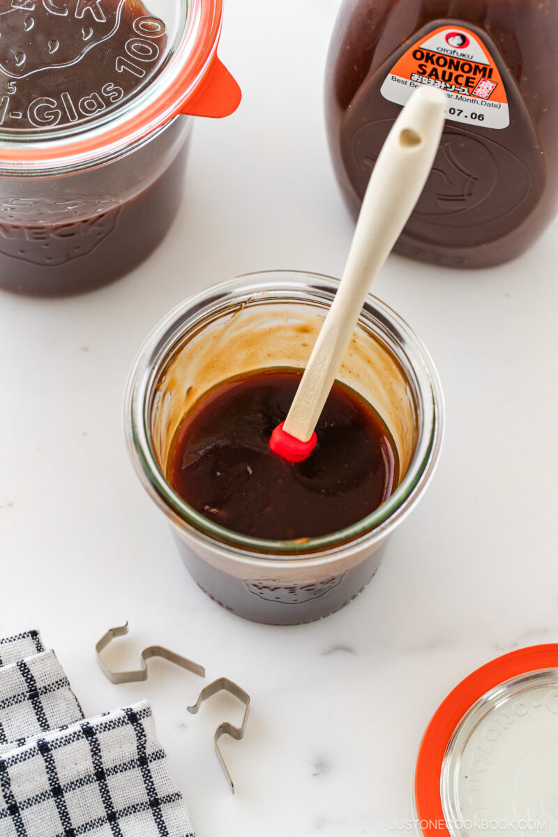 A glass jar filled with dark brown sauce and a small spatula sits on a white surface, surrounded by a sauce bottle, another jar, a jar lid, a metal clip, and a checkered cloth.