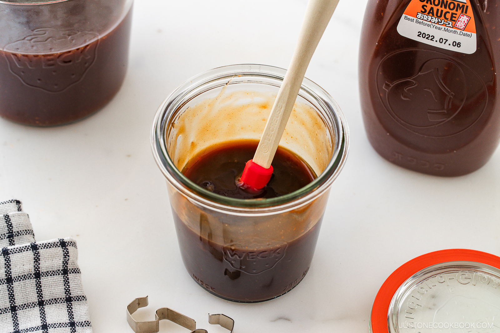 A glass jar filled with a dark brown sauce and a red spatula, surrounded by two bottles of sauce, a jar lid, a checkered cloth, and a metal clip on a white surface.