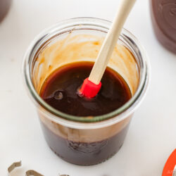 A glass jar filled with thick brown sauce, stirred with a small red and white spatula, sits on a white surface beside a jar lid, a striped cloth, and another jar.