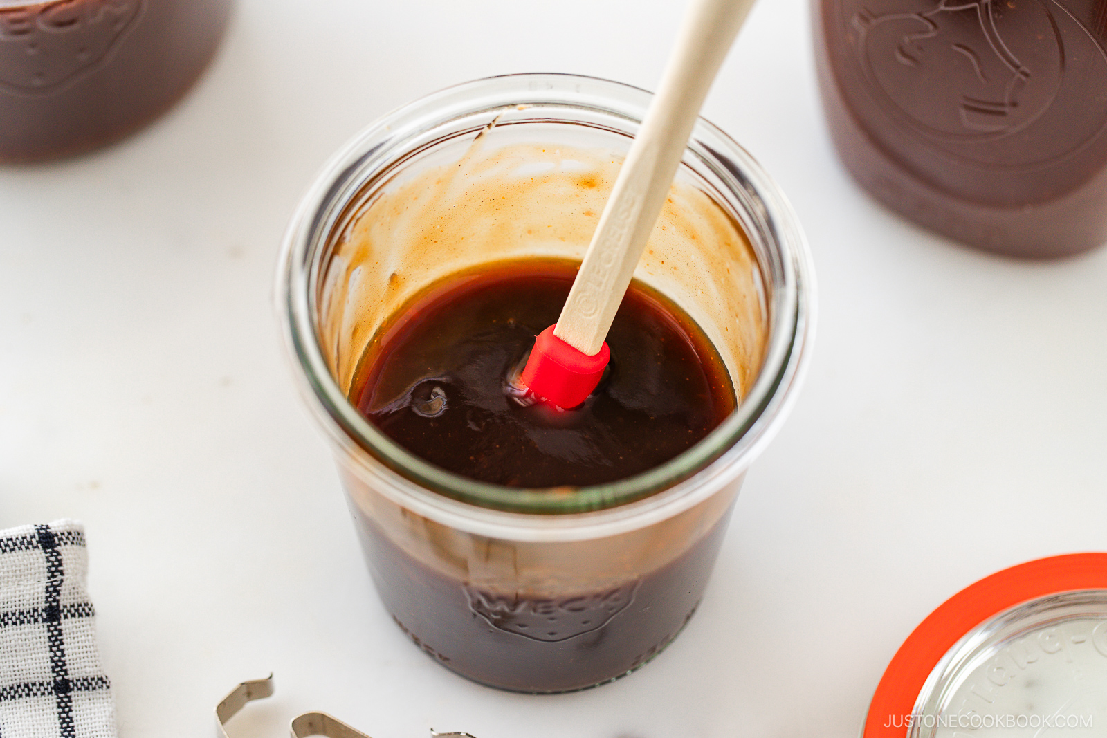 A glass jar filled with thick brown sauce, stirred with a small red and white spatula, sits on a white surface beside a jar lid, a striped cloth, and another jar.