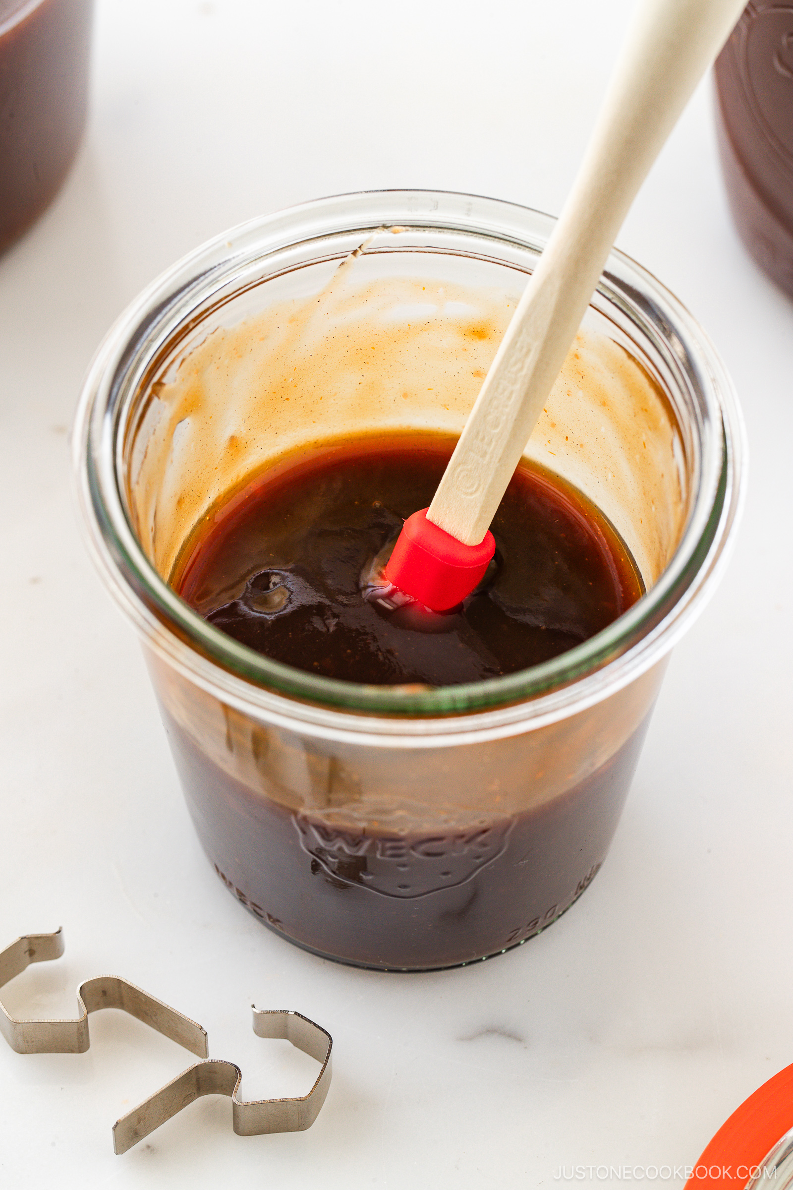 A glass jar filled with dark brown sauce sits on a white surface. A red and white spatula rests inside the jar, and a small metal cookie cutter is nearby. The sauce has a glossy texture.