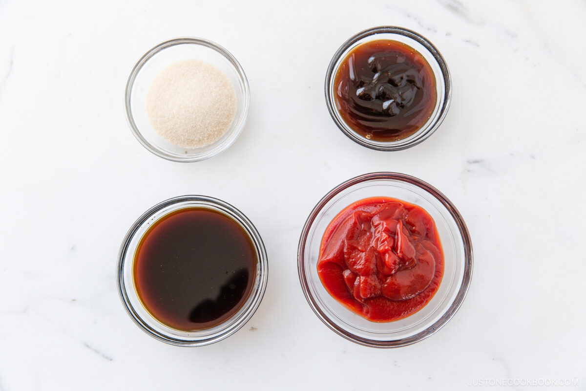 Four small glass bowls on a white surface, each containing a different ingredient: granulated sugar, a thick dark brown sauce, a dark liquid (likely soy sauce), and a red paste or sauce (possibly ketchup).