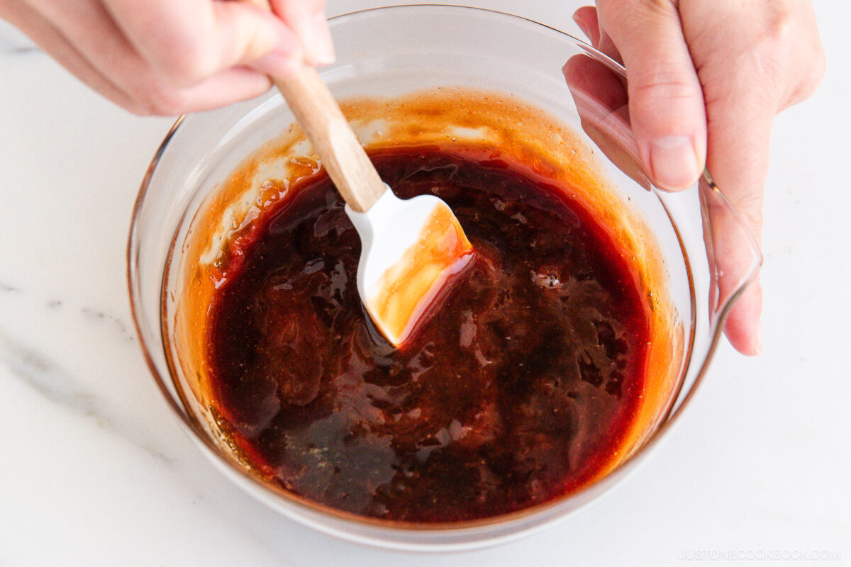 A person stirs a thick, reddish-brown sauce in a glass bowl with a white spatula on a white surface.