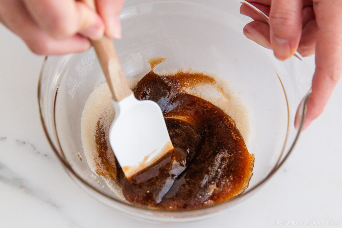 Close-up of hands using a spatula to mix dark brown liquid and granulated sugar in a clear glass bowl on a white surface.