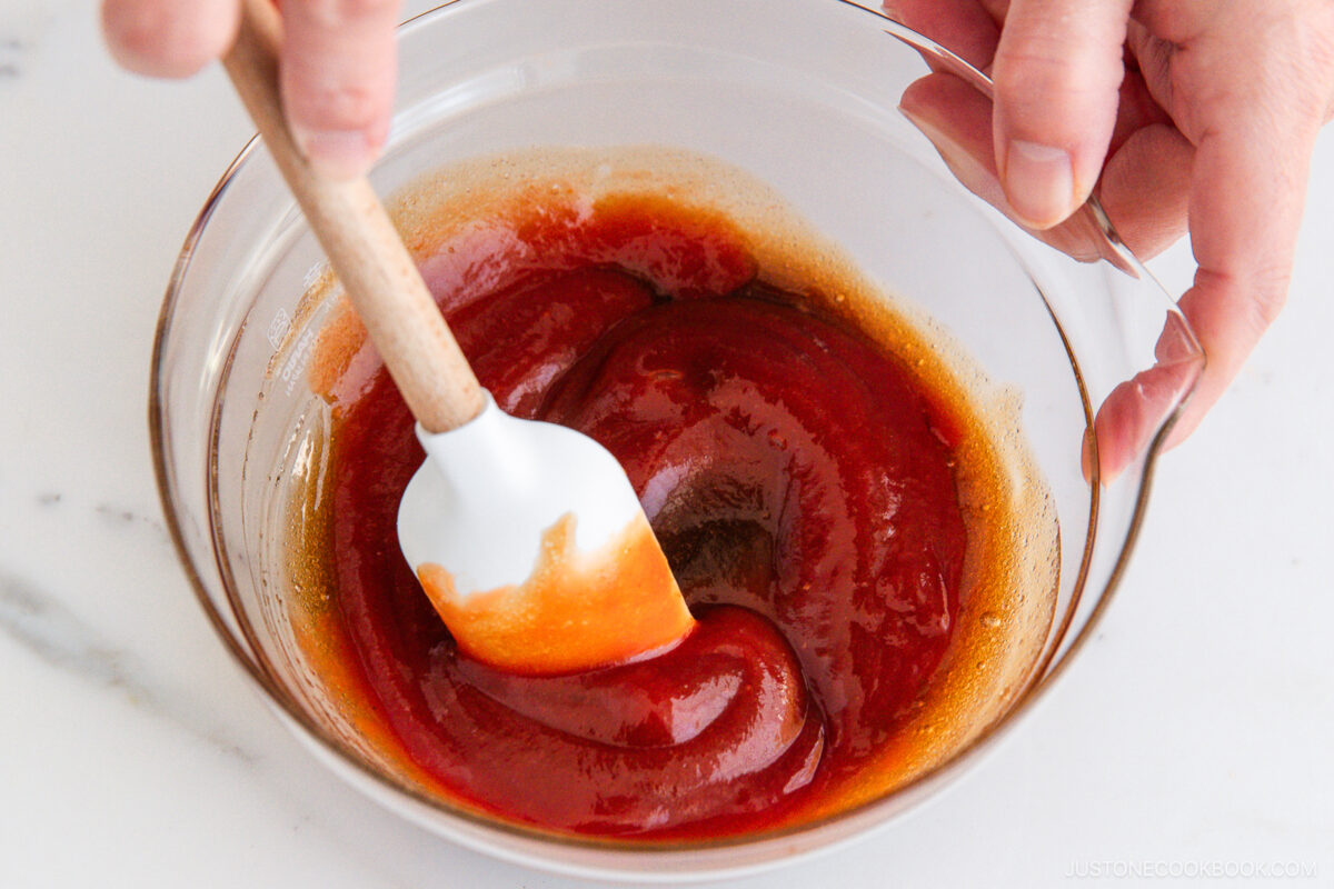 A person uses a white spatula to mix a thick, red sauce in a clear glass bowl on a white surface.