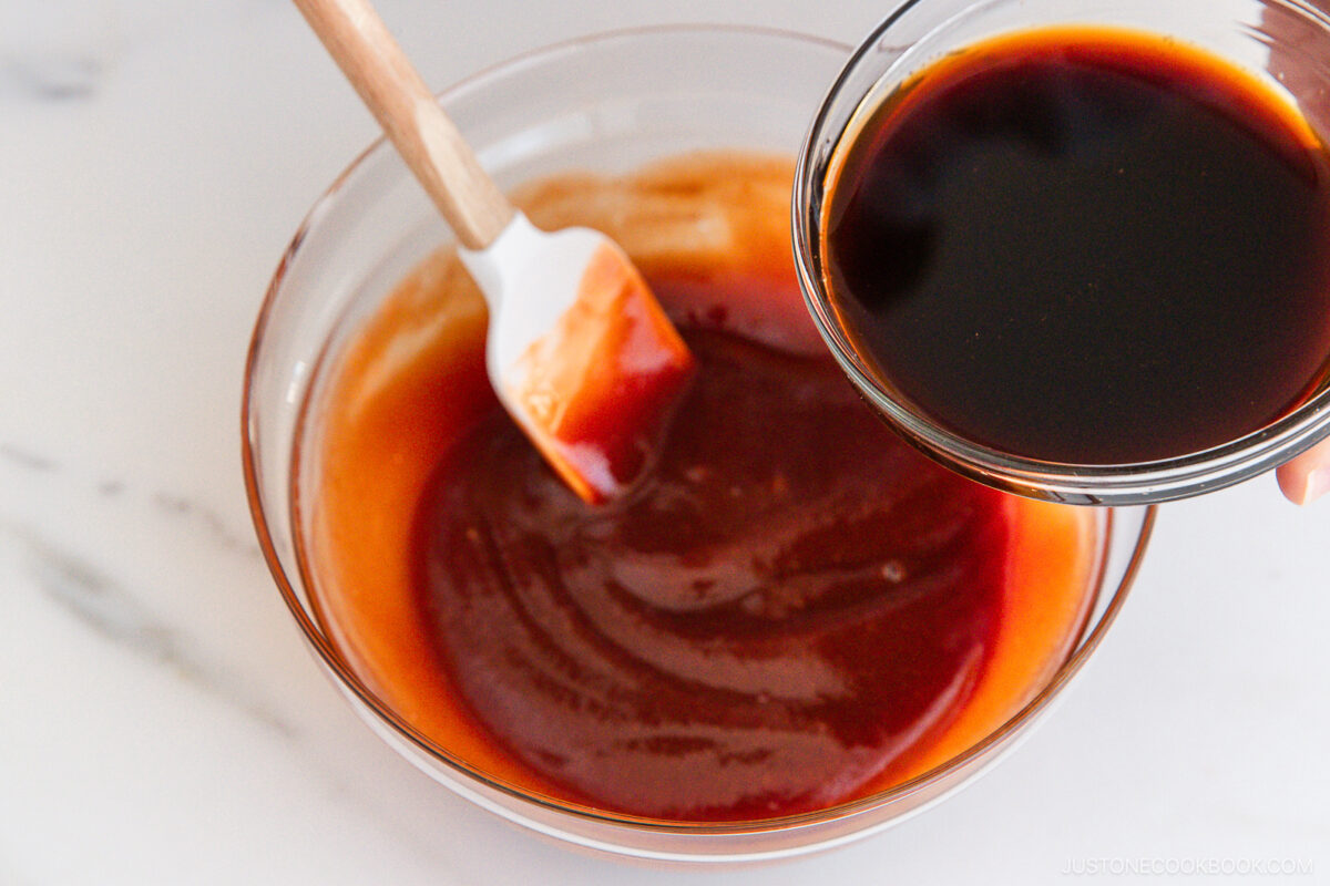 A glass bowl with a red-brown sauce being mixed with a white spatula, while another hand holds a small bowl of dark liquid above, ready to pour it in.