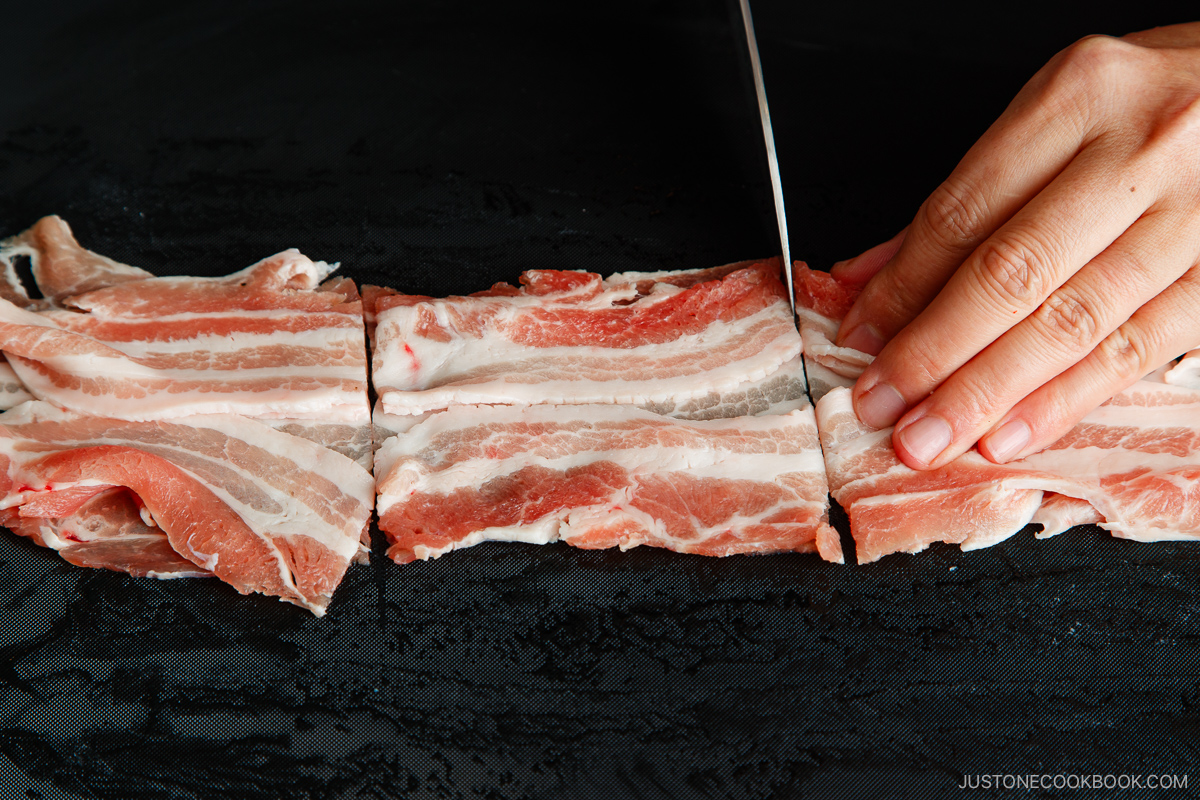 A hand slices raw pork belly strips into sections on a black cutting board with a sharp knife.