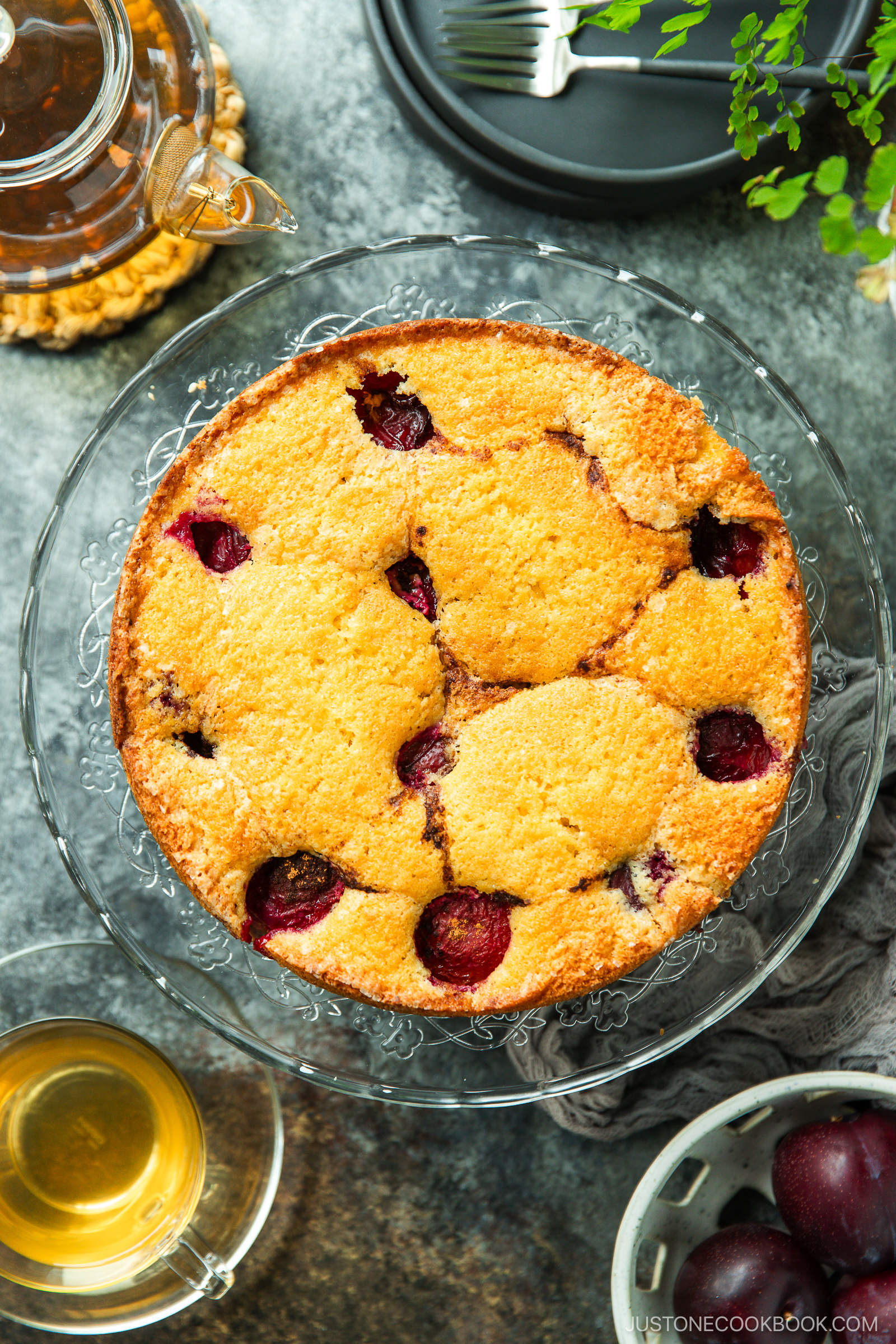 A round cherry cake with a golden-brown top is displayed on a glass cake stand. Nearby are a cup of tea, a teapot, a plate with a fork, and a green plant, all on a gray tabletop.