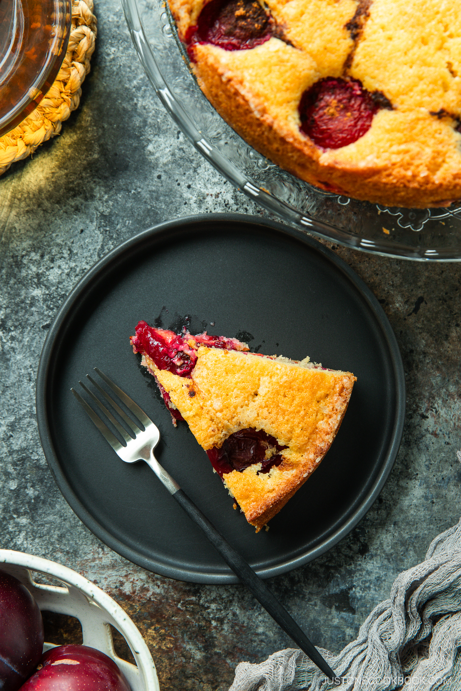 A slice of golden plum cake sits on a black plate with a fork beside it. The rest of the cake, decorated with baked plum halves, is on a glass stand nearby. Fresh plums are in a basket at the bottom of the image.