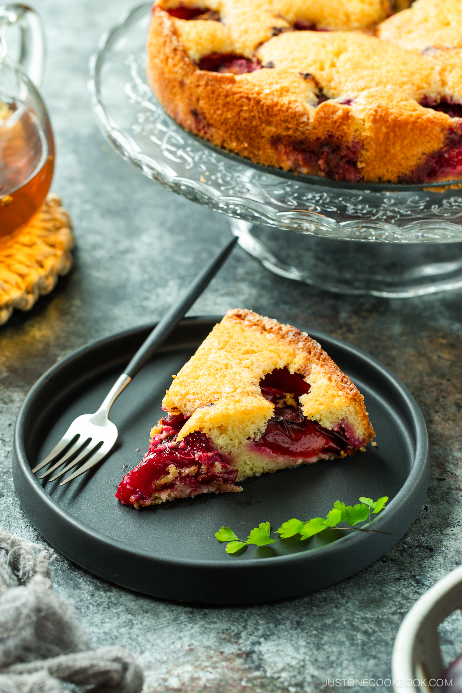 A slice of plum cake sits on a black plate with a fork, garnished with a small green herb, while the rest of the cake is displayed on a glass stand in the background.