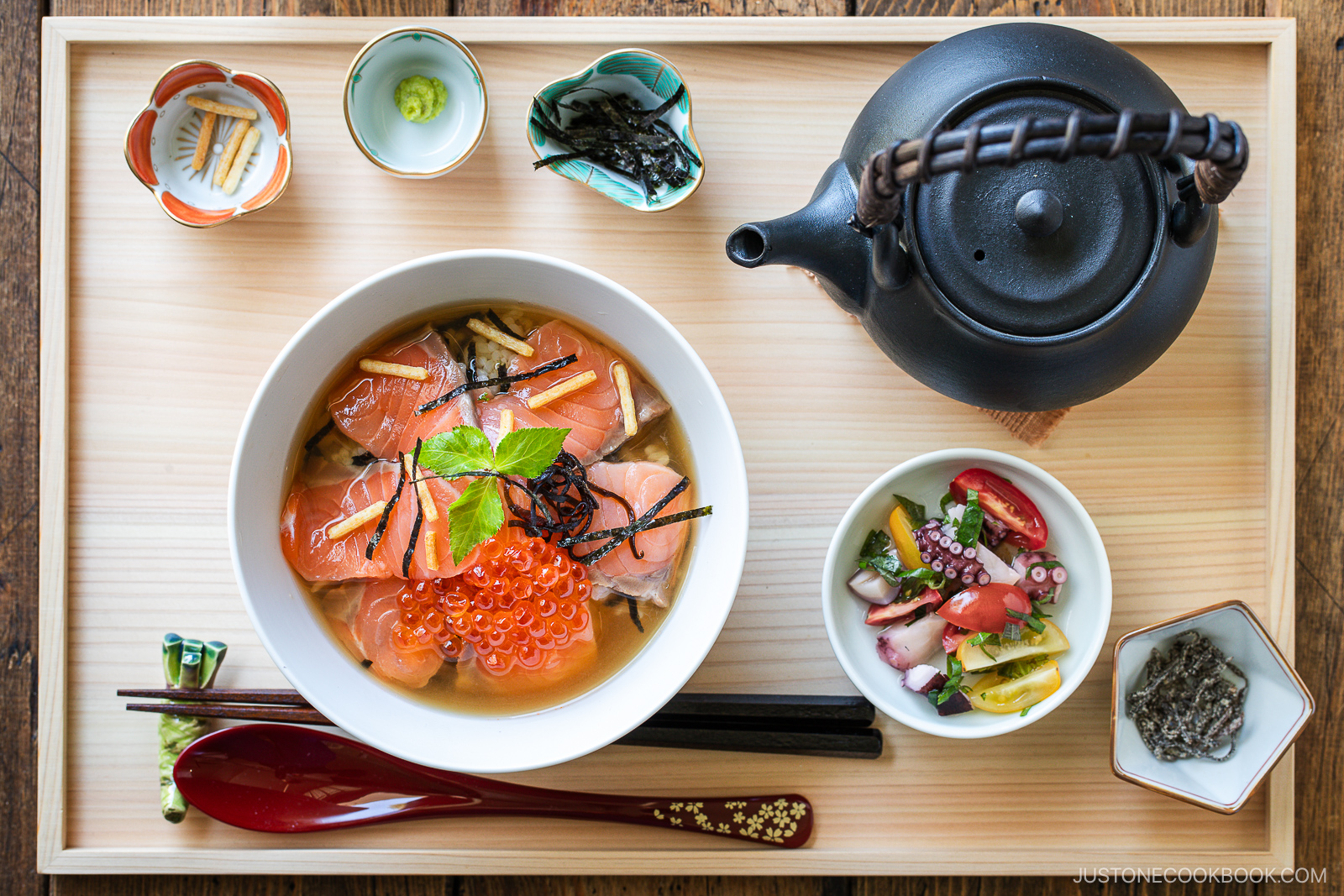 A tray with a bowl of rice topped with salmon slices, ikura (salmon roe), and garnishes, a side of pickled vegetables and seafood, small dishes of condiments, a teapot, and utensils on a wooden tray.