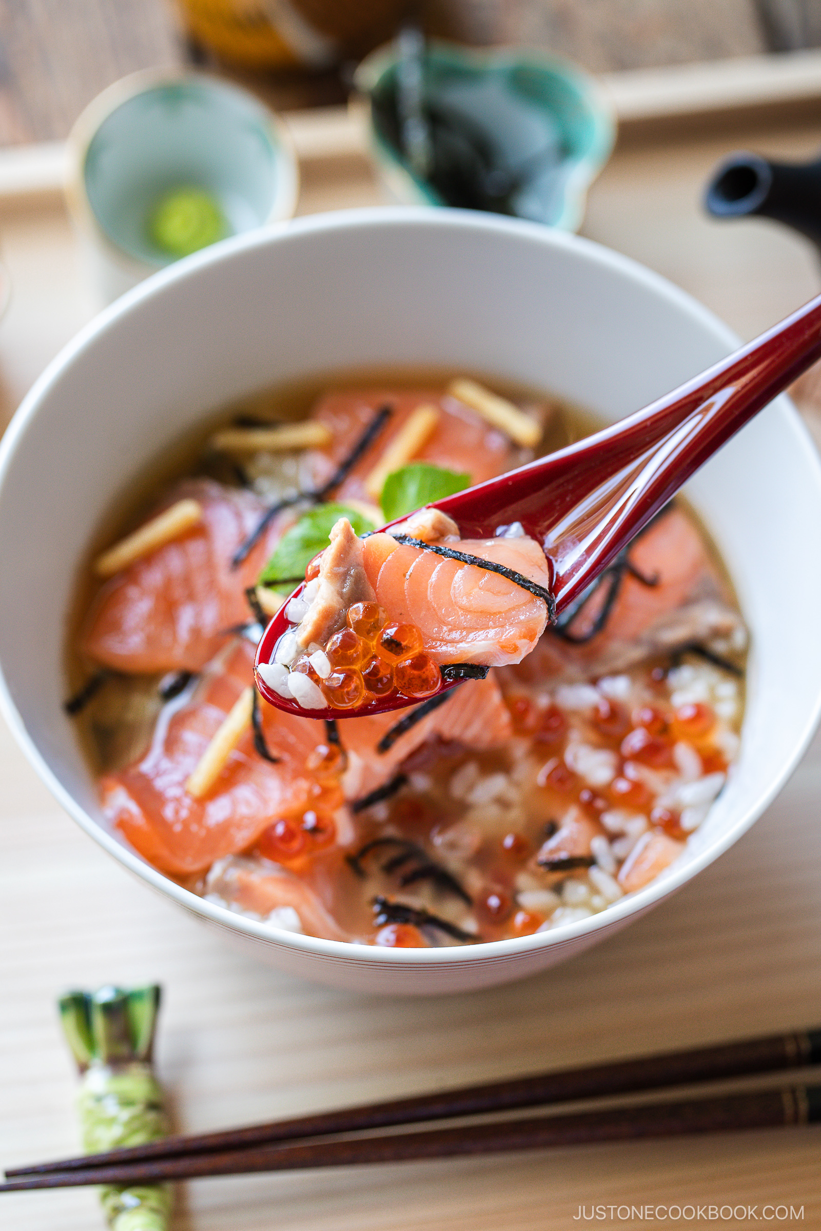 A close-up of a red spoon holding a bite of salmon sashimi, rice, and fish roe above a bowl of Japanese soup with more salmon, rice, and garnishes. Chopsticks and wasabi are visible on the side.