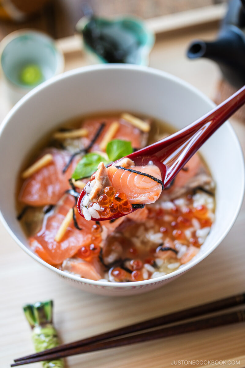 A bowl of Japanese soup topped with raw salmon slices, salmon roe, and seaweed. Red chopsticks hold a piece of salmon above the bowl. A wasabi dollop and chopstick rest are on the wooden table.