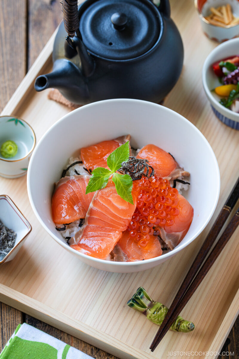 A bowl of rice topped with salmon sashimi, salmon roe, and garnished with green herbs sits on a wooden tray next to a black teapot, pickles, wasabi, and chopsticks.