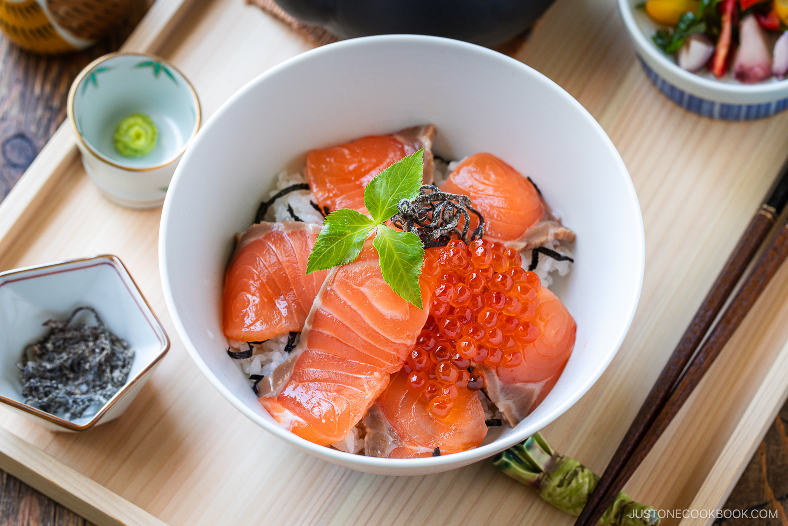 A bowl of Japanese rice topped with slices of raw salmon, salmon roe, and a green garnish. The bowl is on a wooden tray with small dishes of wasabi, pickles, and other side items.