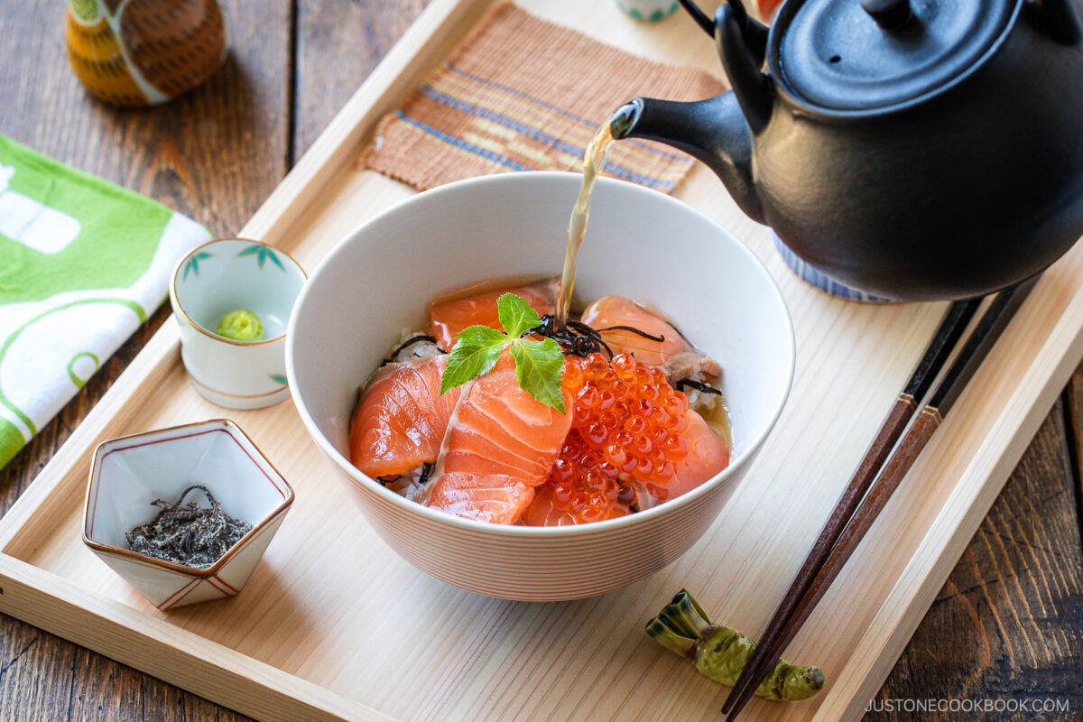 A bowl of Japanese rice topped with salmon sashimi, salmon roe, and herbs is being served with hot tea poured from a teapot. Chopsticks and condiments are arranged on a wooden tray beside the bowl.