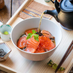 A bowl of Japanese rice topped with salmon sashimi, salmon roe, and herbs is being served with hot tea poured from a teapot. Chopsticks and condiments are arranged on a wooden tray beside the bowl.