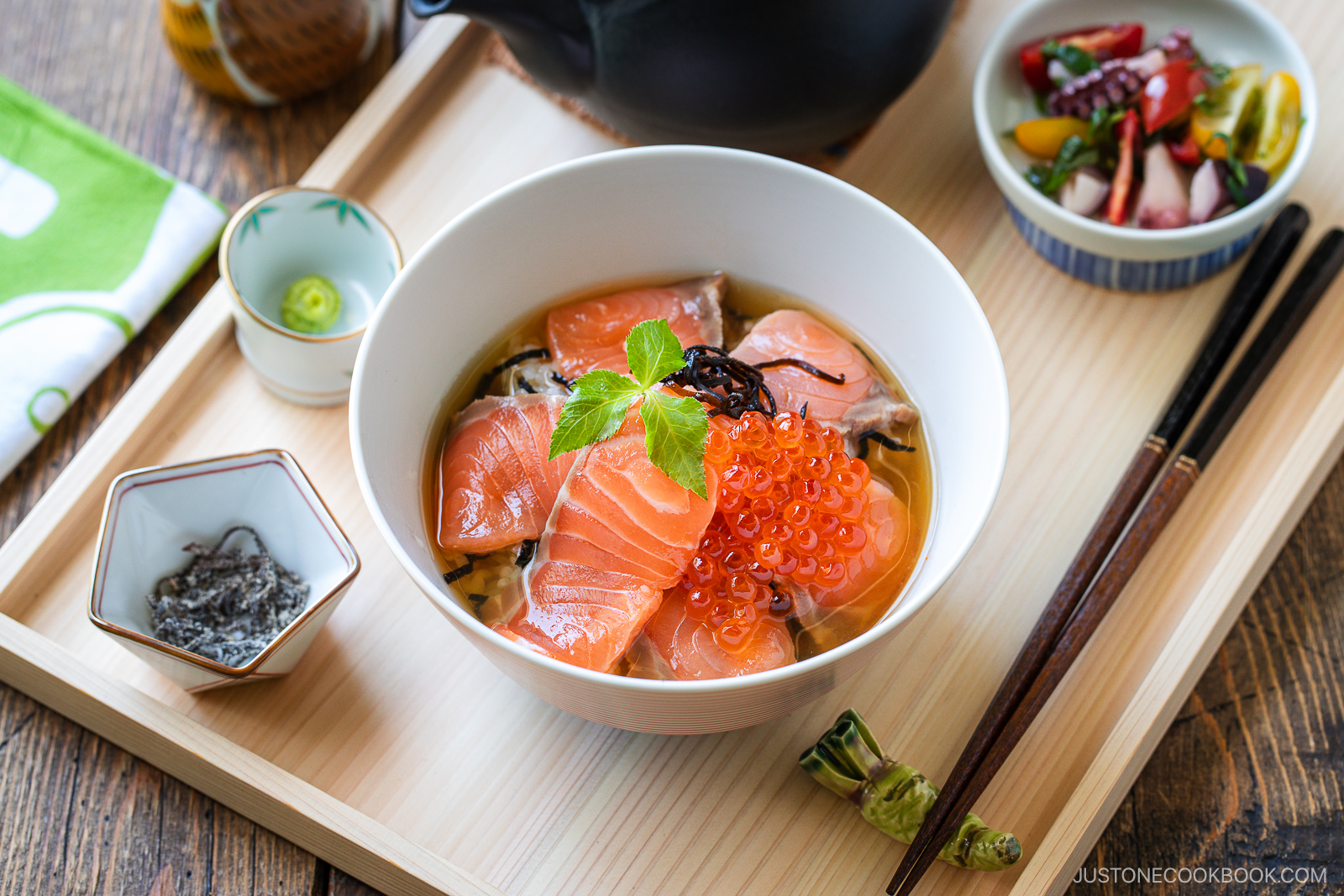 A bowl of Japanese seafood rice topped with fresh salmon, salmon roe, and garnished with a green leaf, served on a wooden tray with side dishes, chopsticks, and wasabi.