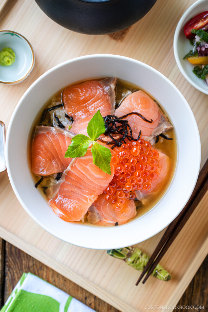 A bowl of Japanese rice topped with raw salmon slices, salmon roe, seaweed, and herbs. Small dishes with wasabi and pickled vegetables, chopsticks, and a teapot are seen on the wooden tray.